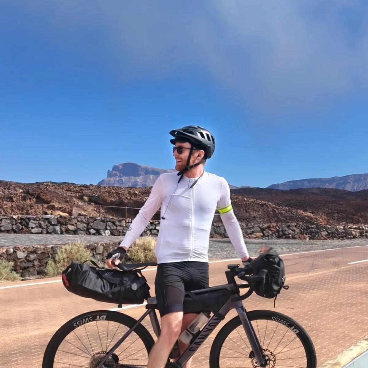 Jonas Acker in a white jersey and helmet, standing with a loaded bikepacking bike on a mountain road under a blue sky.